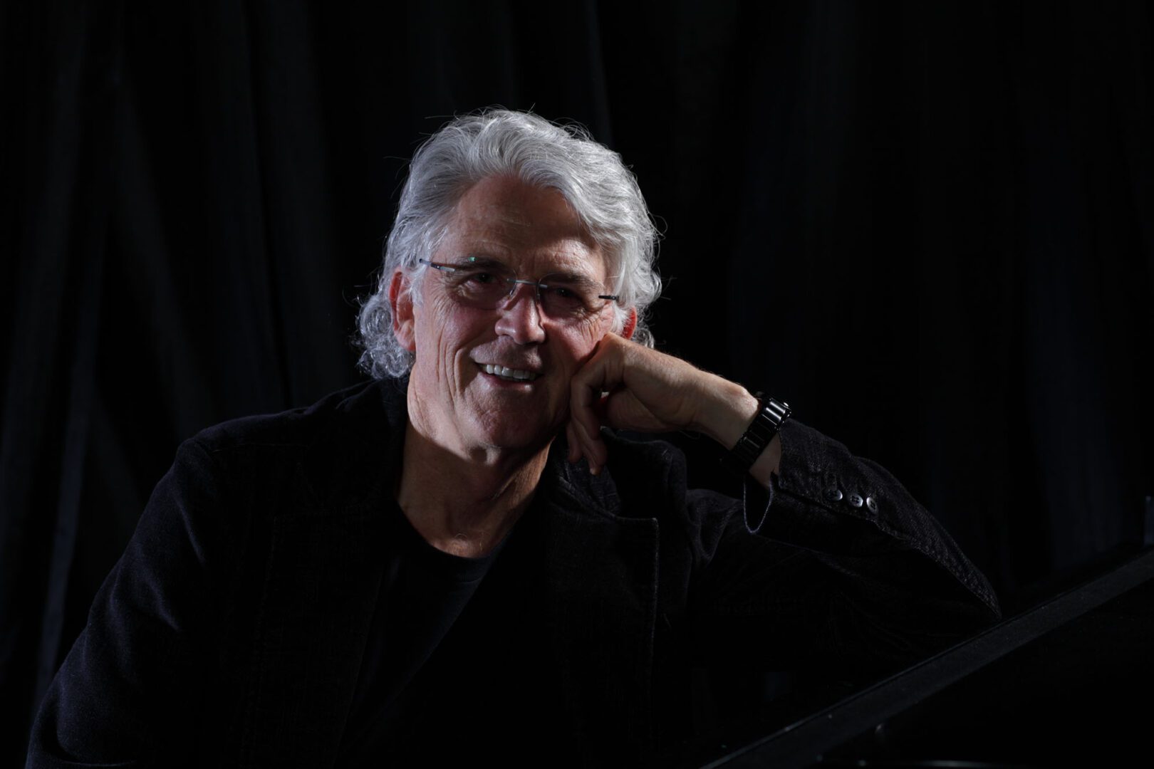 A man with white hair and glasses sitting in front of a piano.